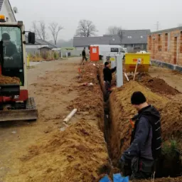 Wykopany rów w ziemi, widoczni pracownicy w trakcie pracy, jeden z nich montuje białą skrzynkę elektryczną w rowie, obok stoi minikoparka i w tle budowa domu.