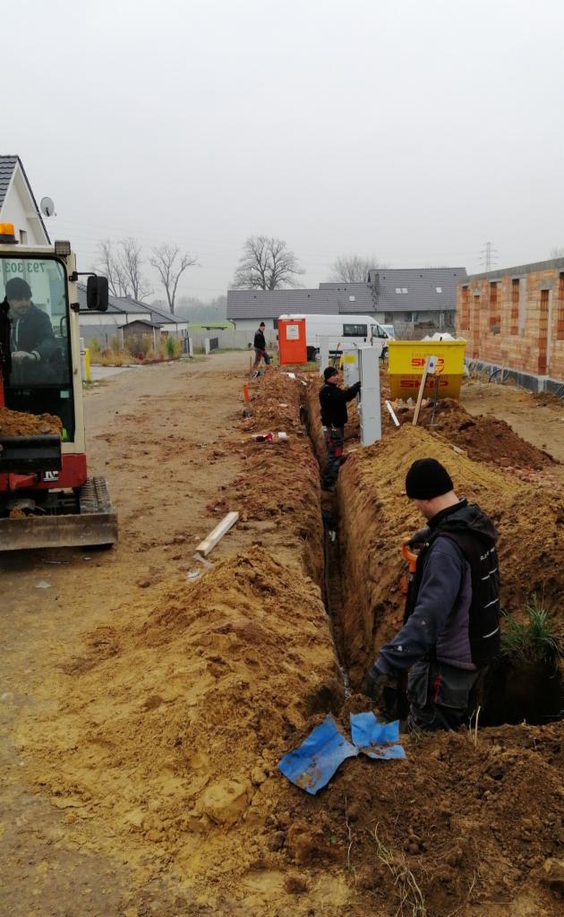 Wykopany rów w ziemi, widoczni pracownicy w trakcie pracy, jeden z nich montuje białą skrzynkę elektryczną w rowie, obok stoi minikoparka i w tle budowa domu.