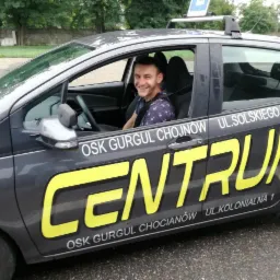Uśmiechnięty instruktor siedzi w samochodzie oznaczonym logo 'CENTRUM OSK GURGUL', Chojn&oacute;w, ul. Solskiego 31, ul. Kolonialna 1, z charakterystycznym znakiem 'L' na dachu.