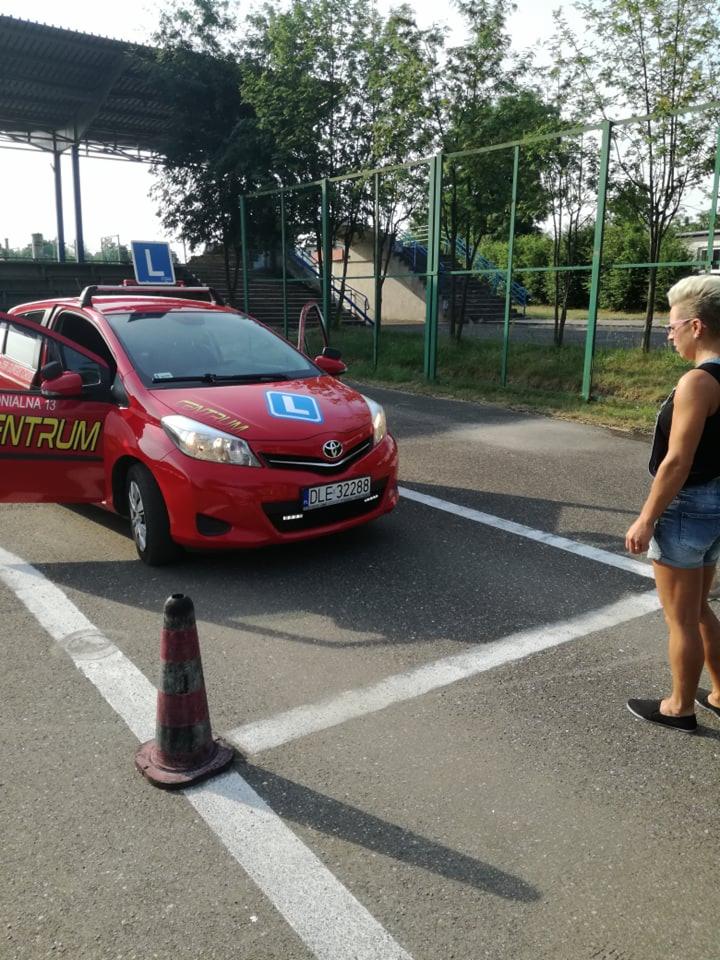 Czerwony samochód z oznaczeniem 'L' na dachu i masce, zaparkowany na placu manewrowym z pachołkiem, obok stoi kobieta w dżinsowych szortach.