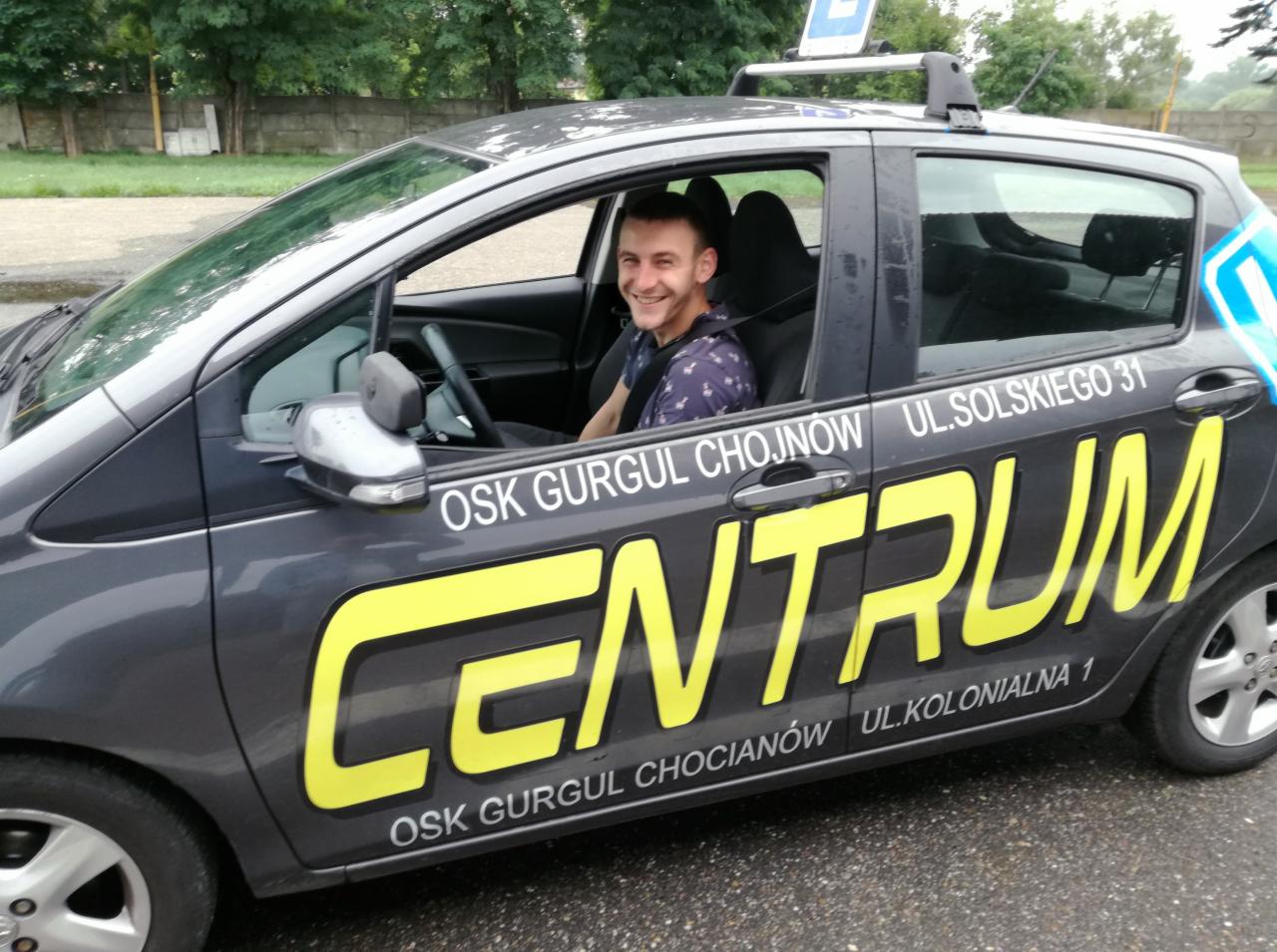 Uśmiechnięty instruktor siedzi w samochodzie oznaczonym logo 'CENTRUM OSK GURGUL', Chojnów, ul. Solskiego 31, ul. Kolonialna 1, z charakterystycznym znakiem 'L' na dachu.