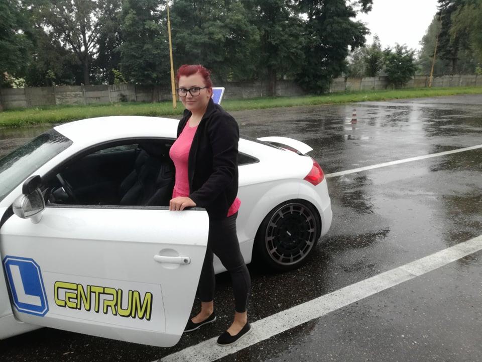 Instruktorka w okularach, opierająca się o białe Audi TT z naklejką szkoły jazdy 'L CENTRUM' na drzwiach, na mokrym placu manewrowym.