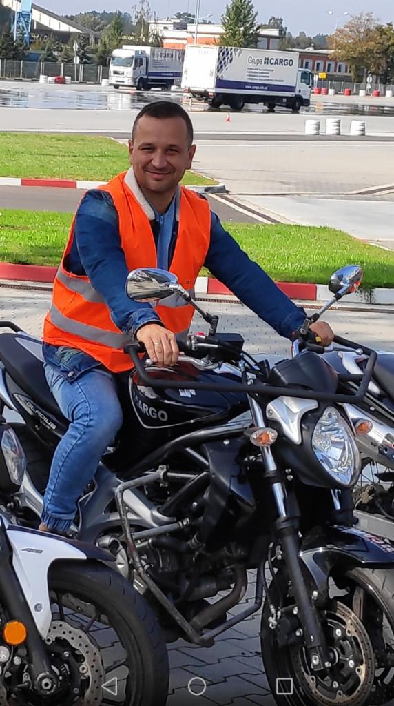 Instruktor w pomarańczowej kamizelce siedzi na czarnym motocyklu Suzuki Gladius, w tle widoczny budynek i ciężarówka z napisem Grupa CARGO.