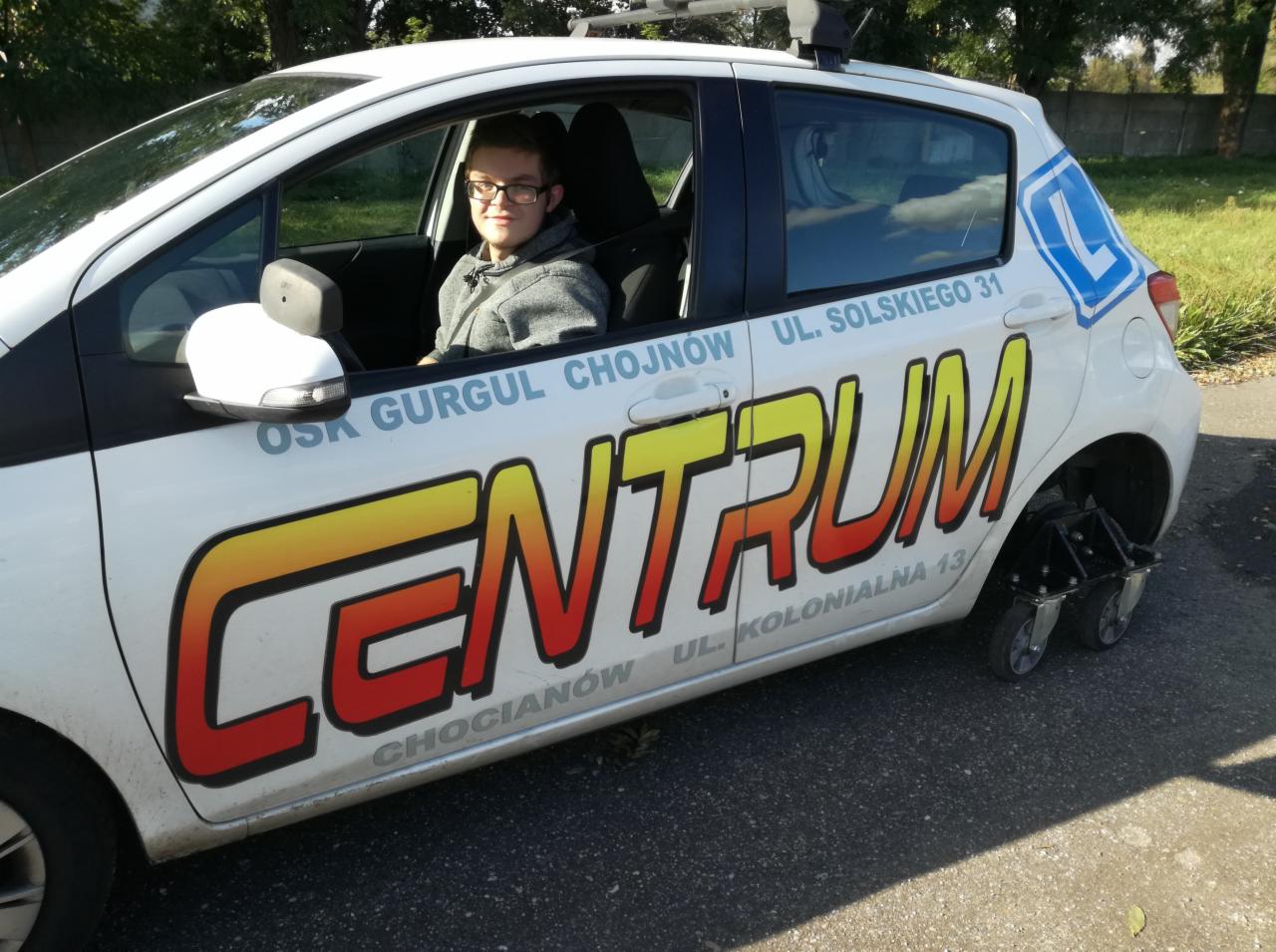 Biały samochód z naklejką 'CENTRUM' i adresem, dodatkowe małe kółka z tyłu, młody mężczyzna w okularach siedzi za kierownicą.