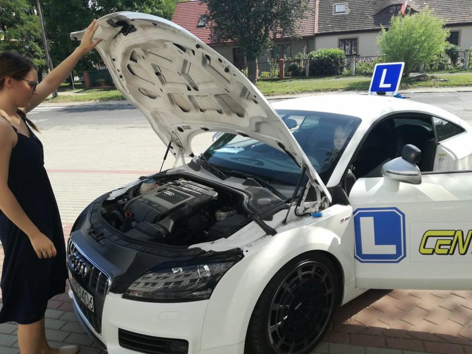 Kobieta w sukience otwiera maskę białego Audi TT z oznaczeniami szkoły jazdy, widoczny silnik i znak 'L' na dachu.