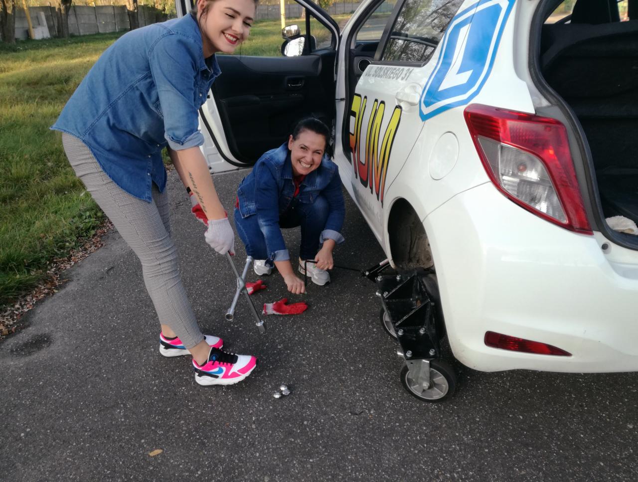 Dwie uśmiechnięte kobiety przy białym samochodzie oznaczonym literą 'L', jedna wymienia koło przy użyciu klucza, druga pomaga, widoczny podnośnik pod autem.
