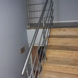 Schody drewniane z nowoczesną balustradą ze stali nierdzewnej, proste metalowe pręty, szara ściana z dekoracją.