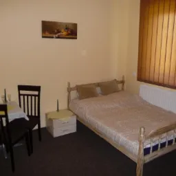 Pokój hotelowy z drewnianym łóżkiem, stolikiem z szampanem i dwoma krzesłami, oraz obrazem martwej natury na ścianie w ciepłych barwach.