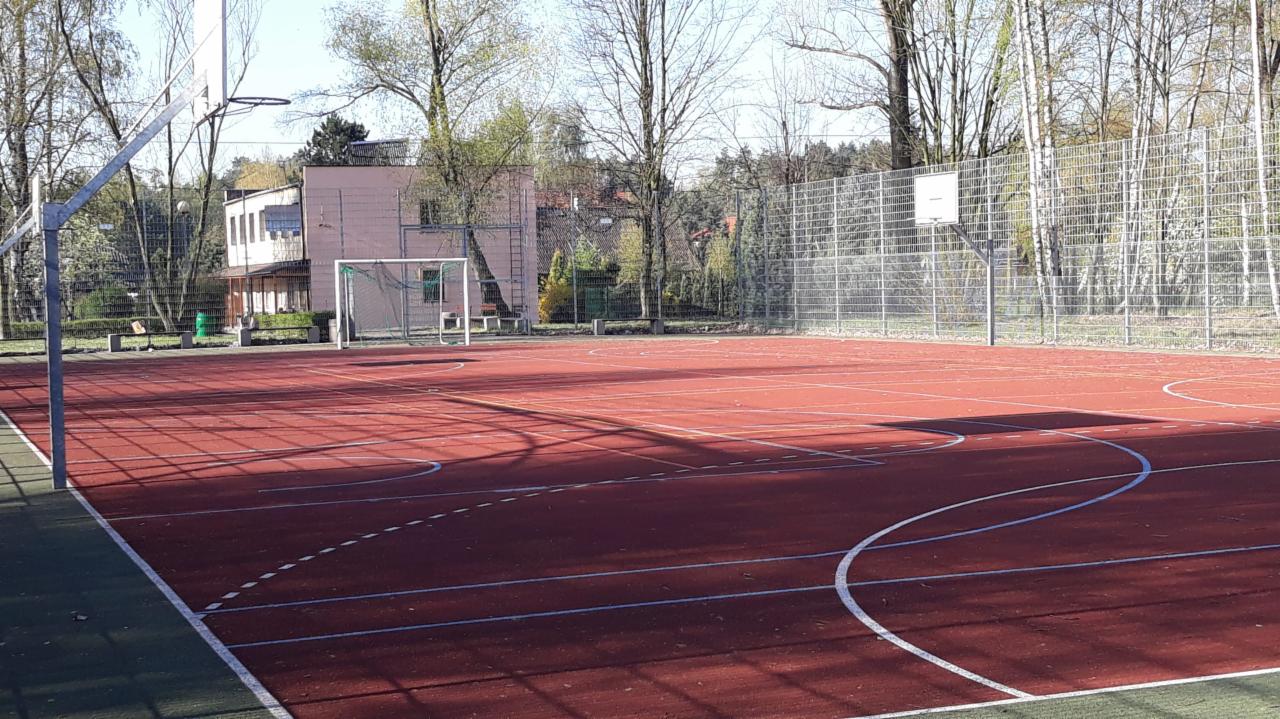 Czerwone boisko wielofunkcyjne z bramką do piłki nożnej i koszem do koszykówki, otoczone siatką i drzewami, z budynkiem w tle.