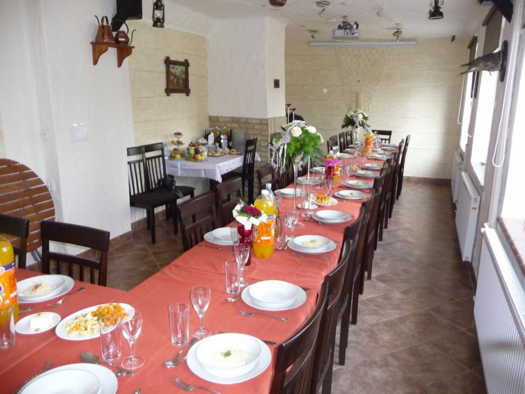 Długi stół nakryty czerwonym obrusem, zastawiony talerzami, sztućcami i szklankami, z dekoracjami kwiatowymi i napojami, przygotowany dla dużej grupy osób w jasnym pomieszczeniu z brązową podłogą.