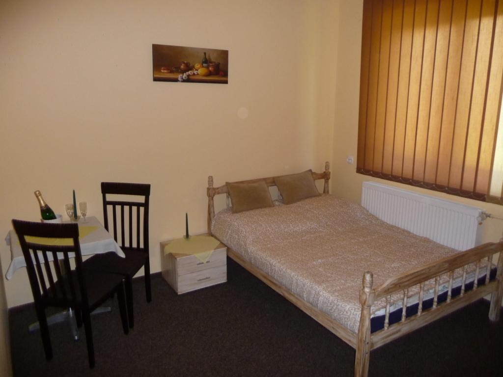 Pokój hotelowy z drewnianym łóżkiem, stolikiem z szampanem i dwoma krzesłami, oraz obrazem martwej natury na ścianie w ciepłych barwach.