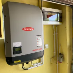 Szary falownik Fronius Symo zamontowany na żółtej ścianie w garażu, obok skrzynka z bezpiecznikami i przewody w białych rurkach PCV.