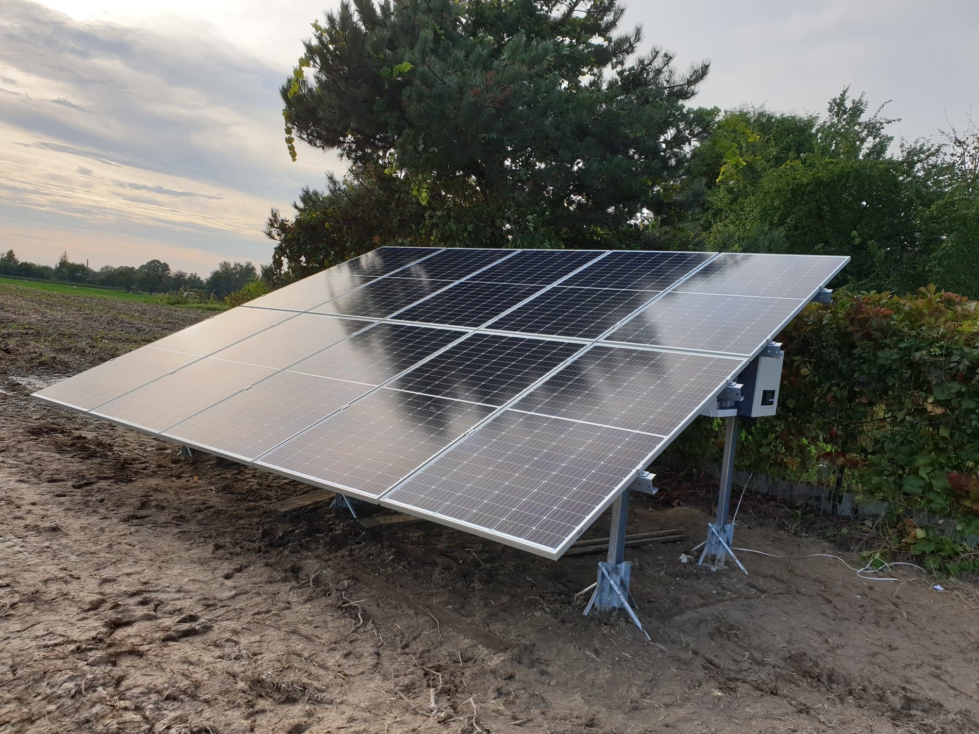 Zestaw paneli fotowoltaicznych na metalowej konstrukcji, zamontowany na gruncie, z widocznym inwerterem i okablowaniem, na tle drzew i pochmurnego nieba.