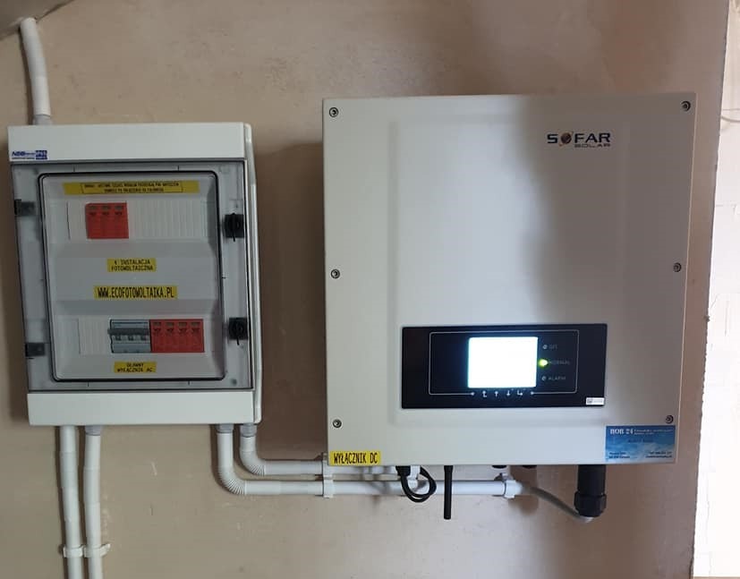 Biała skrzynka rozdzielcza z widoczną instalacją fotowoltaiczną i adresem strony internetowej, obok falownik Sofar Solar z wyświetlaczem i naklejką informacyjną, rury instalacyjne prowadzone po...
