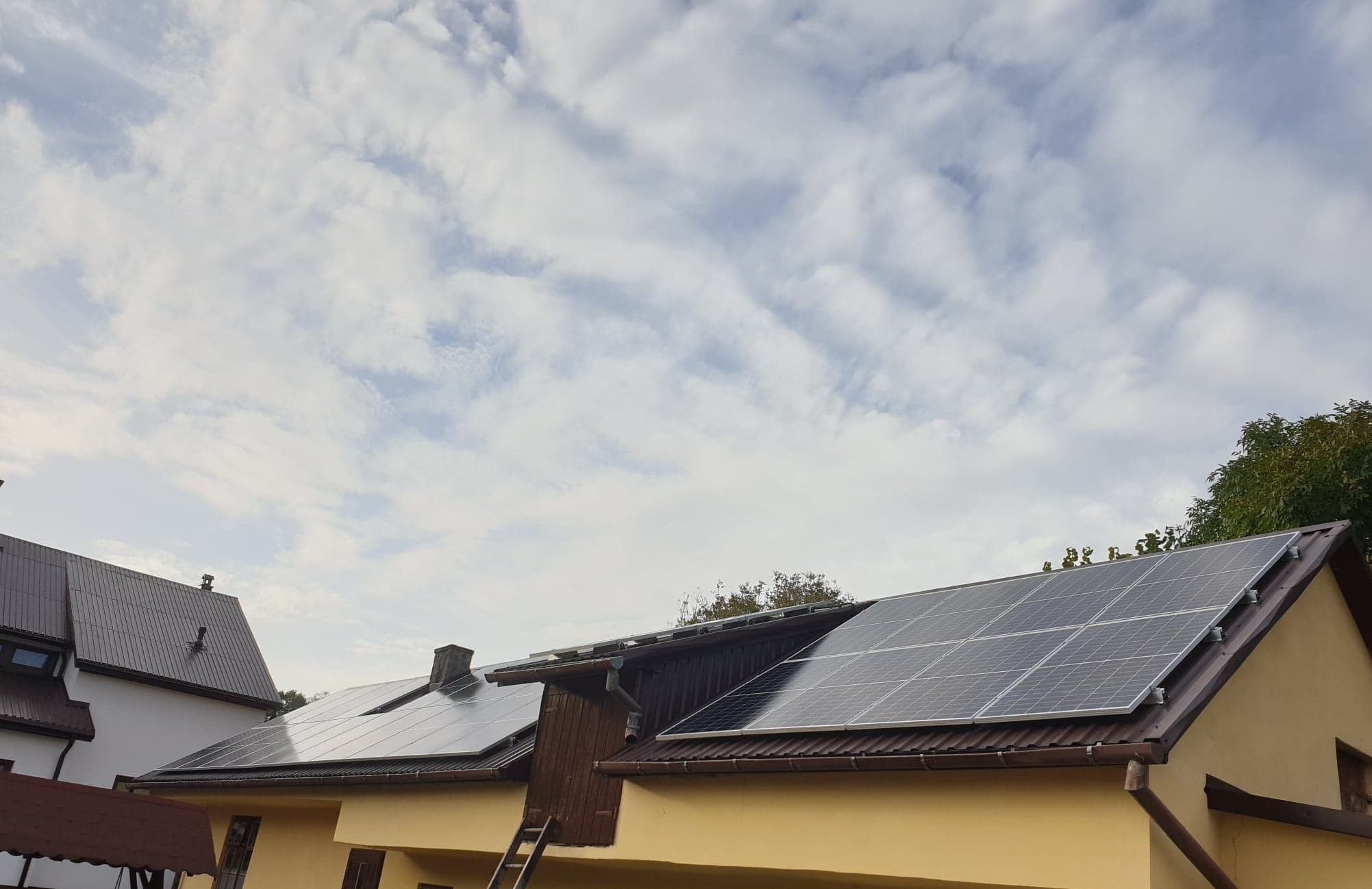 Panele fotowoltaiczne zamontowane na dachu dwuspadowym domu z żółtą elewacją i brązowym dachem, częściowo widoczna drabina oparta o ścianę, pochmurne niebo w tle.
