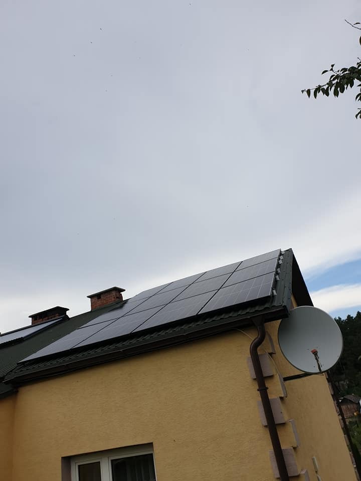 Żółty dom z panelami słonecznymi na dachu, widoczne kominy i antena satelitarna, pochmurne niebo.