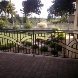 Czarna metalowa balustrada balkonowa z dekoracyjnym motywem fal, na pierwszym planie kostka brukowa, w tle ogród z drewnianym płotem i wiszące kwiaty.