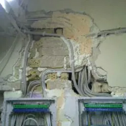 Odsłonięte przewody elektryczne w ścianie z uszkodzonym tynkiem, widoczne puszki rozdzielcze z listwami zaciskowymi.