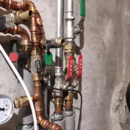 Fragment instalacji grzewczej z miedzianymi rurami, zaworami z czerwonymi i zielonymi uchwytami, manometrem i izolacją na tle surowej, betonowej ściany.