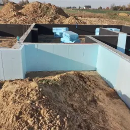 Budowa fundamentów: widok na wykop z izolacją termiczną ścian fundamentowych i zabezpieczeniem hydroizolacyjnym, z hałdami ziemi wewnątrz i na zewnątrz.