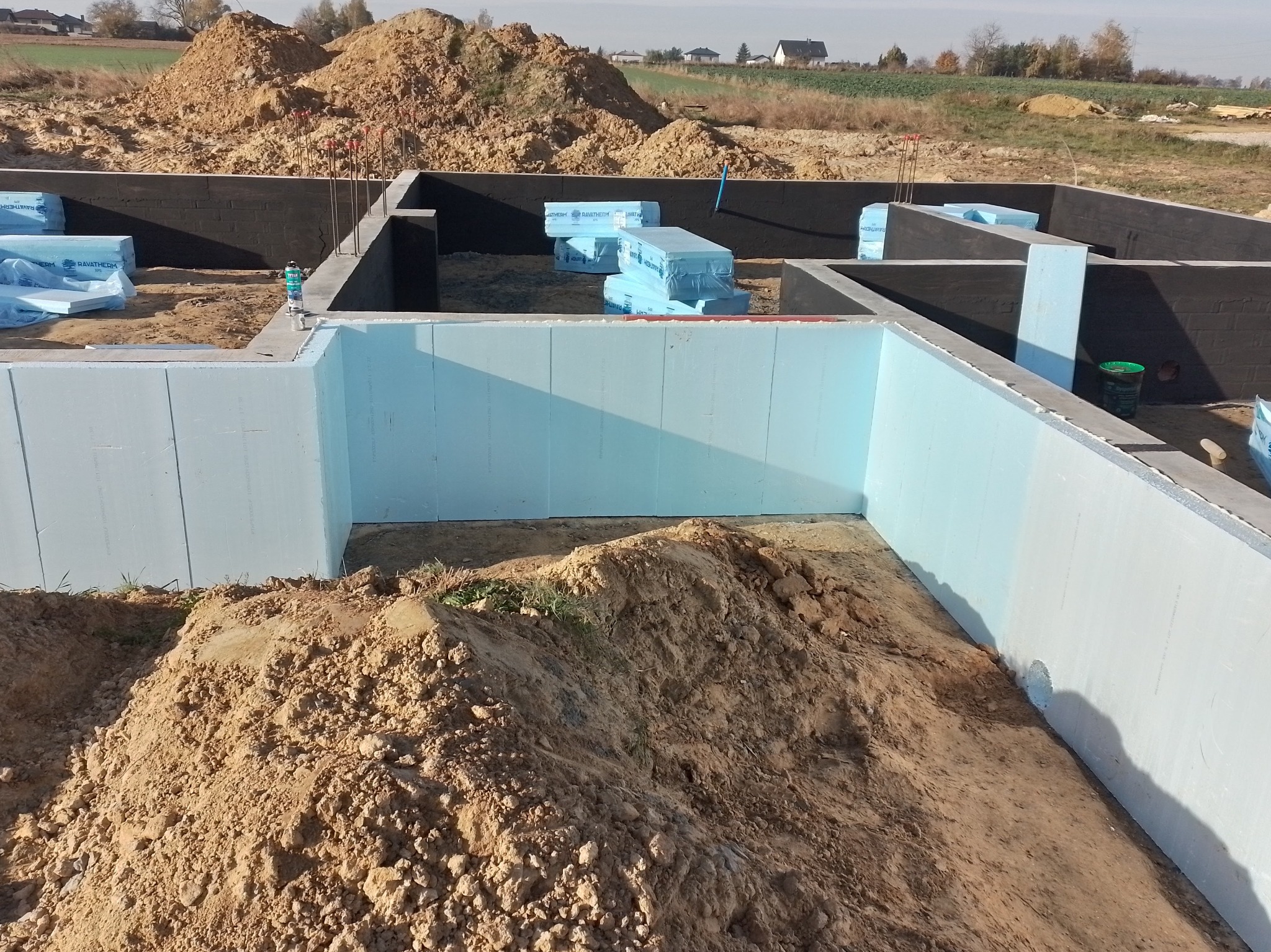 Budowa fundamentów: widok na wykop z izolacją termiczną ścian fundamentowych i zabezpieczeniem hydroizolacyjnym, z hałdami ziemi wewnątrz i na zewnątrz.