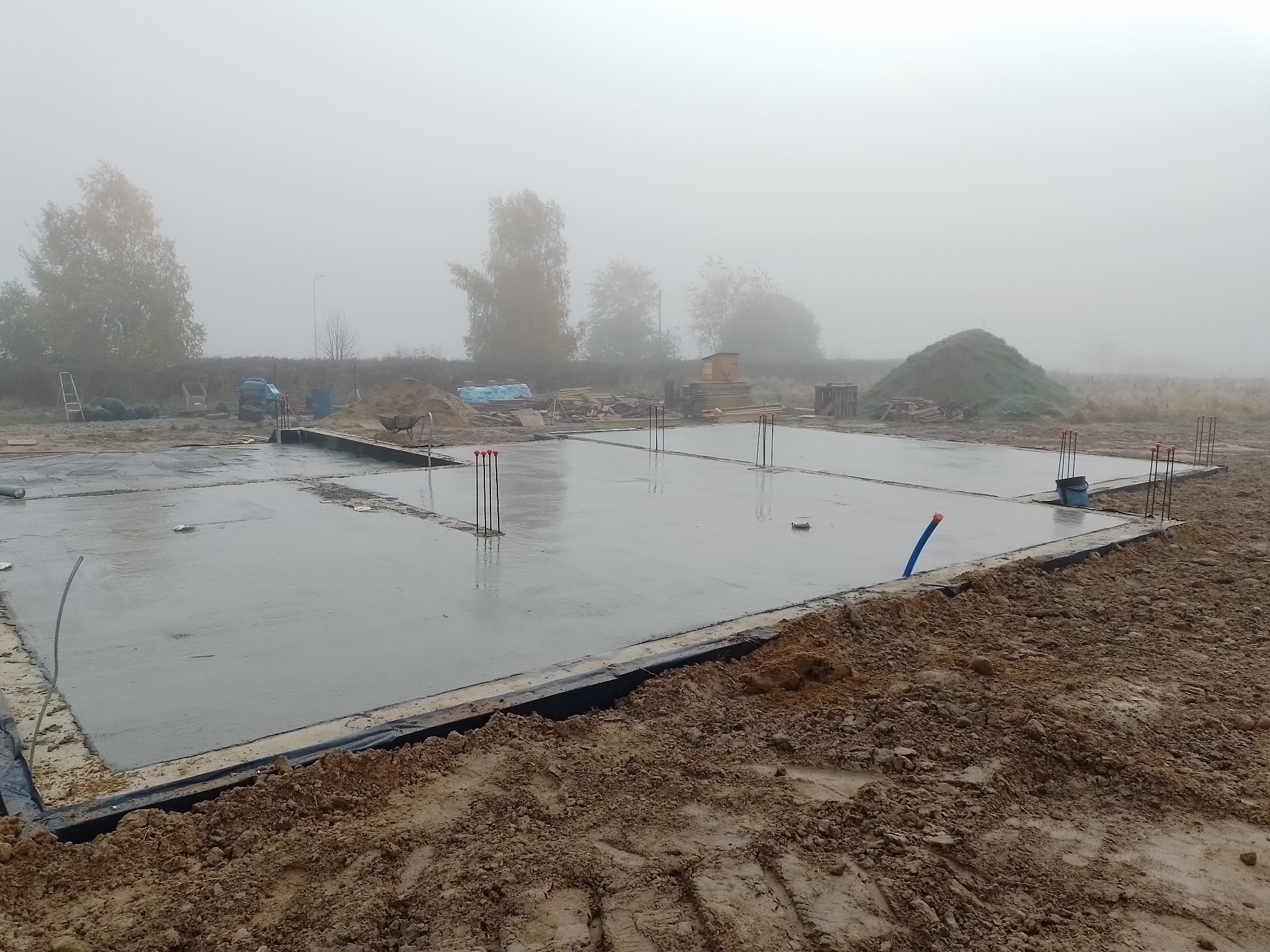 Świeżo wylana, mokra płyta fundamentowa z wystającymi prętami zbrojeniowymi, widoczna w porannej mgle, z przygotowanym gruntem i stertą ziemi w tle.
