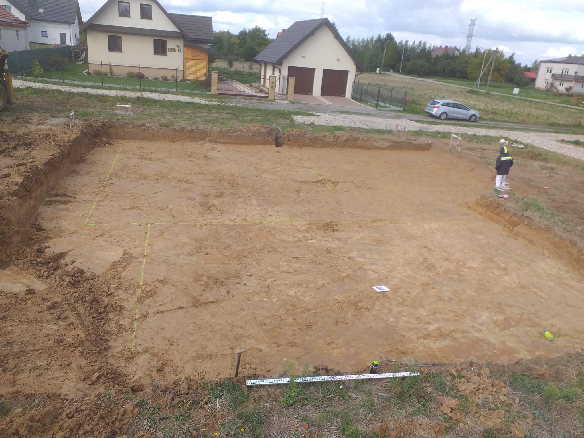 Wykop pod fundamenty domu jednorodzinnego, wytyczone granice budynku, widoczny pracownik w odzieży ochronnej, w tle zabudowa mieszkalna.