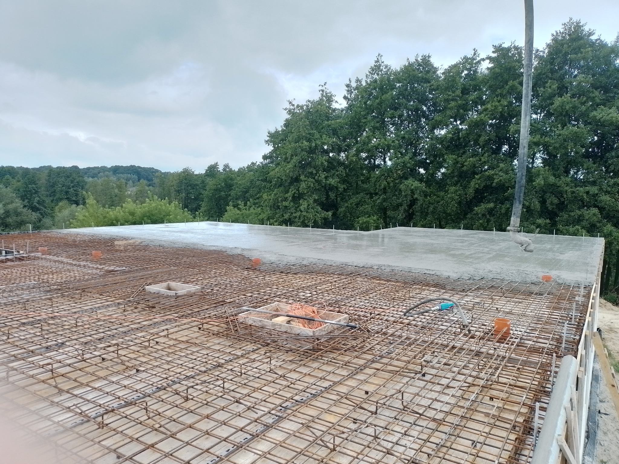 Zalewanie stropu betonem z pompy na siatce zbrojeniowej, widoczne kable elektryczne w korytkach, w tle las.