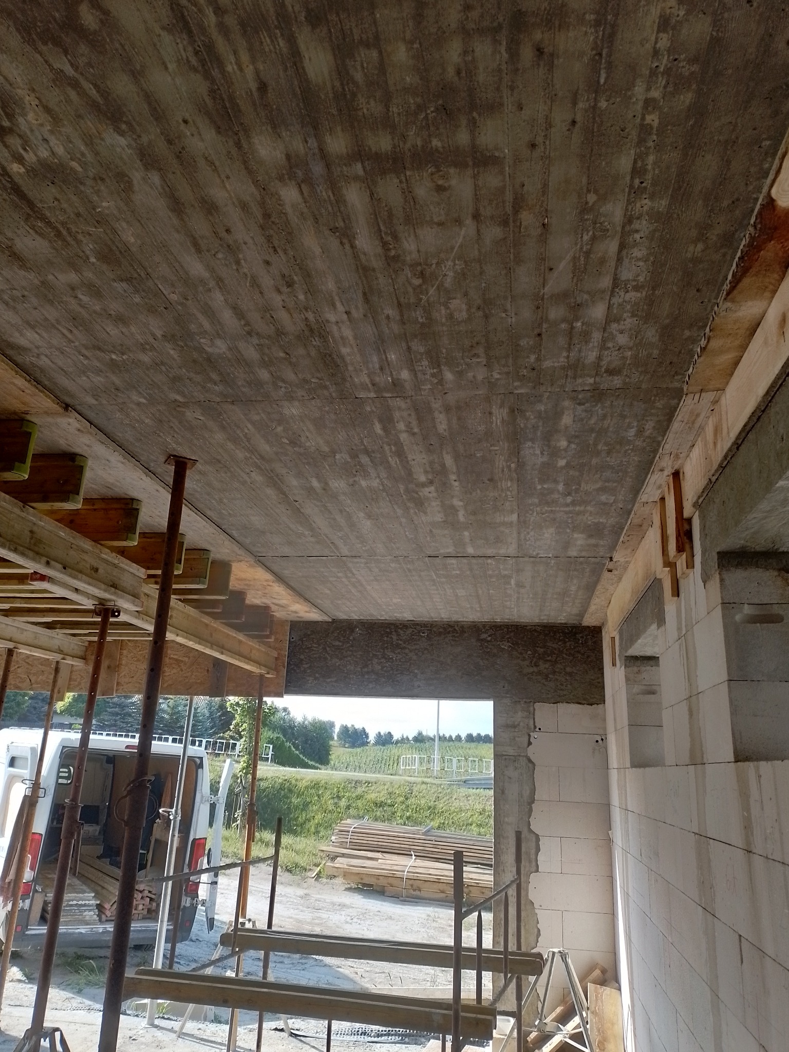 Betonowy strop w trakcie budowy, widoczne stemple podpierające szalunek, ściany z bloczków betonu komórkowego, w tle krajobraz.