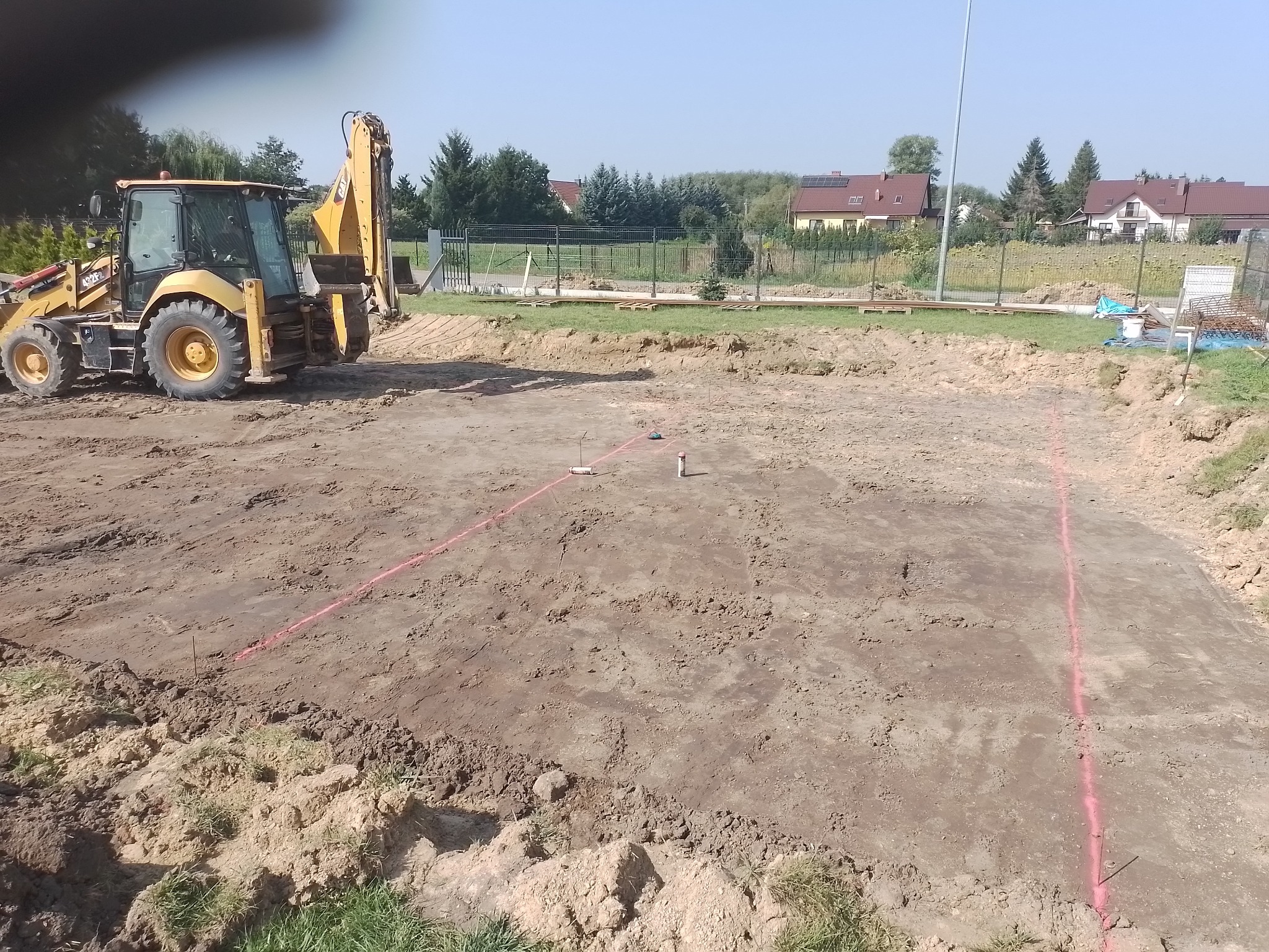 Wyrównany teren pod budowę w Lublinie, wyznaczony różową linią, z koparką w tle i widokiem na domy jednorodzinne.