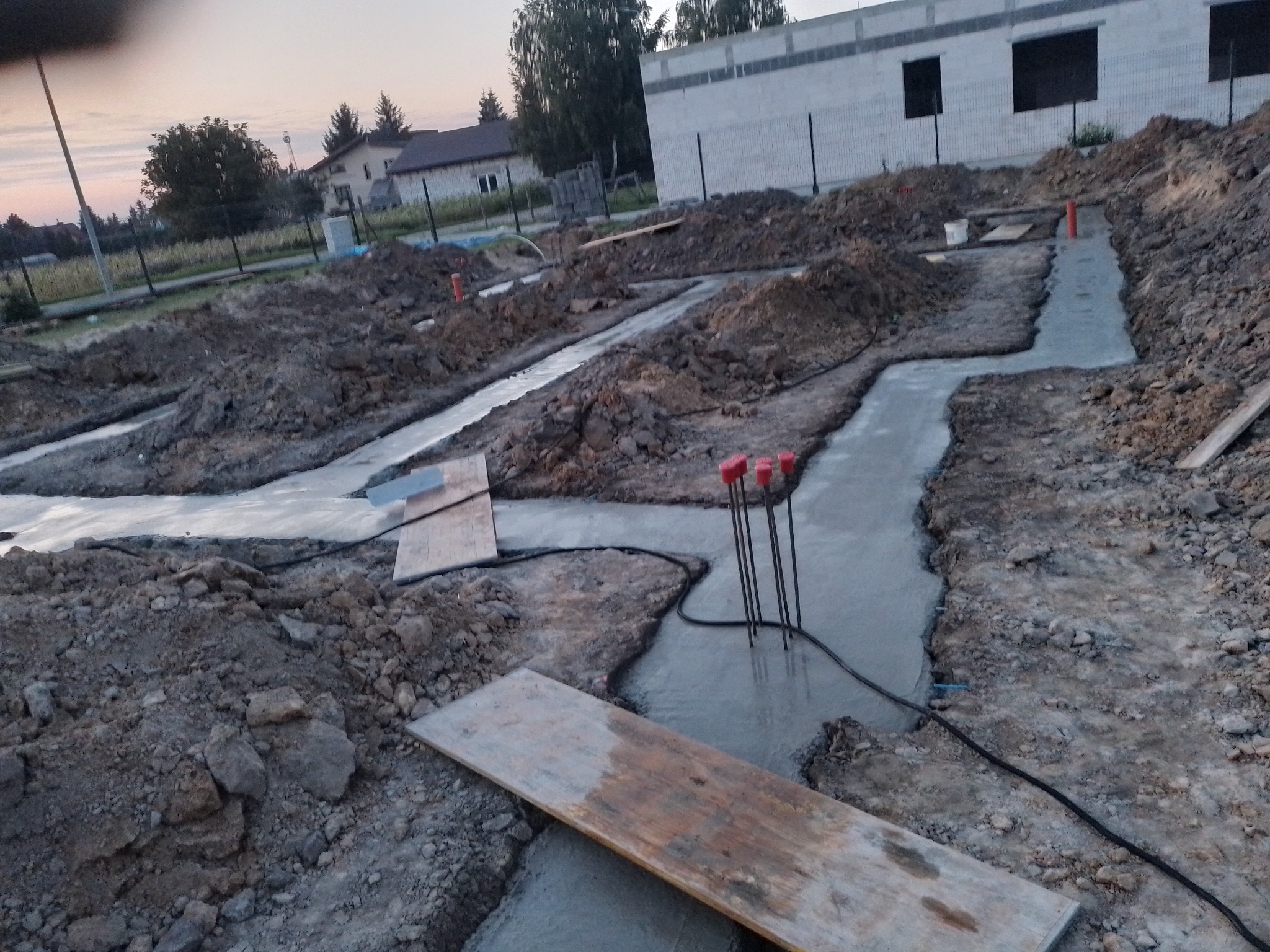 Zalane betonem wykopy pod fundamenty z wystającymi prętami zbrojeniowymi, widoczne deski szalunkowe i folia izolacyjna, w tle budynek w trakcie budowy.