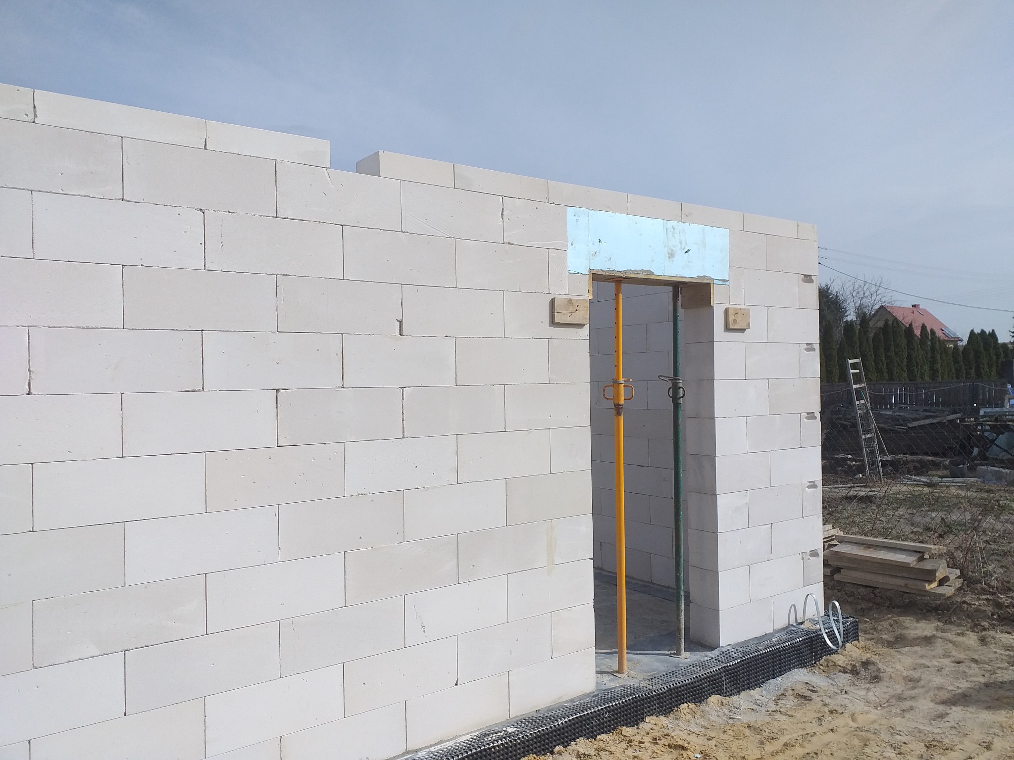 Ściana budynku w trakcie budowy, wzniesiona z białych bloczków, z widocznym otworem drzwiowym podpartym żółtą i zieloną stemplą, z widoczną hydroizolacją fundamentów.