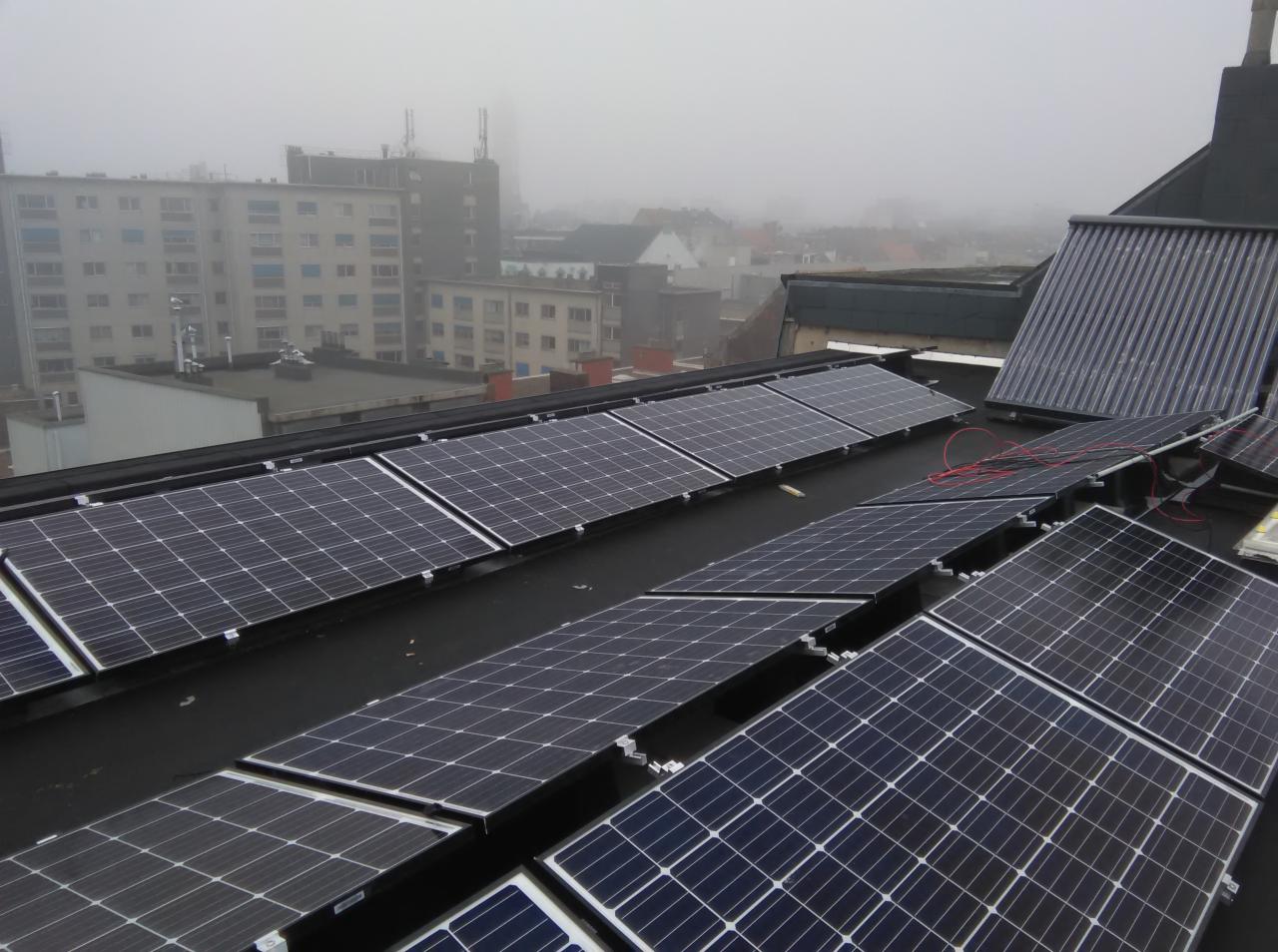 Panele fotowoltaiczne na płaskim dachu budynku w pochmurny dzień, widoczne czerwone przewody, w tle zabudowa miejska spowita mgłą.