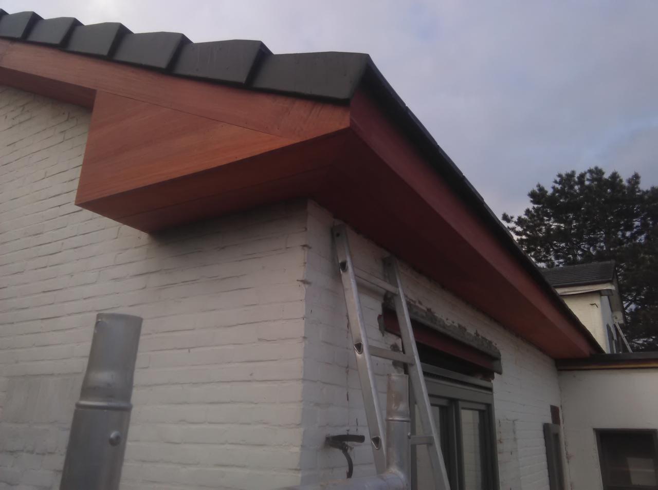 Wykończenie okapu drewnem w kolorze mahoniowym na tle białej, ceglanej ściany budynku, z widoczną aluminiową drabiną opartą o ścianę.