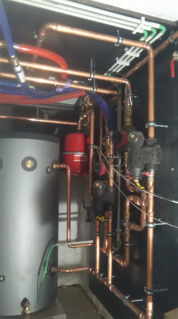 Złożony system rur miedzianych, zbiornik szary, naczynie przeponowe czerwone, pompy obiegowe czarne, rury z tworzyw sztucznych białe i kolorowe, zamontowane w kotłowni na czarnej ścianie.