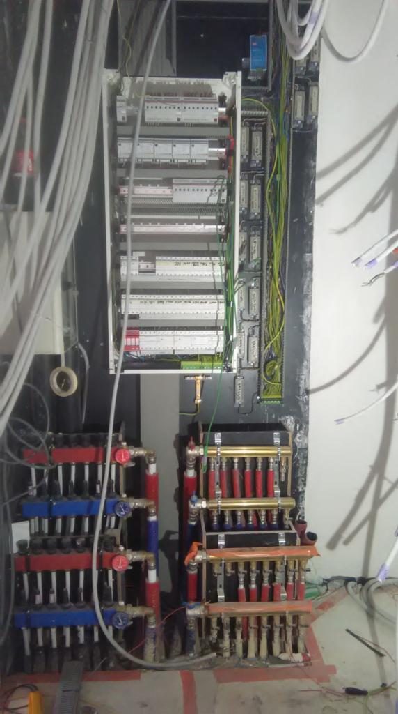 Rozdzielnica elektryczna z licznymi przewodami i bezpiecznikami, obok rozdzielacze instalacji hydraulicznej z rurami w kolorach czerwonym i niebieskim, w trakcie montażu.
