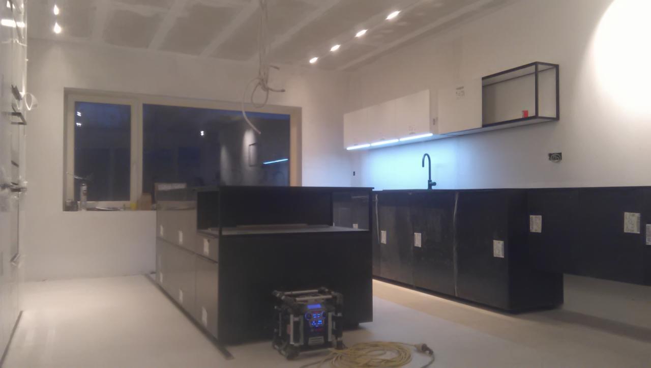 Wnętrze kuchni w trakcie montażu: czarne szafki kuchenne z białymi górnymi szafkami i oświetleniem LED, widoczne niedokończone ściany i sufit z instalacją elektryczną, na podłodze leży zwinięty kabel.