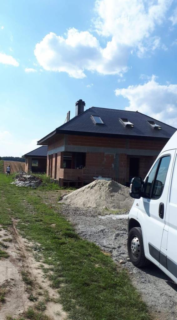 Dwa domy w stanie surowym otwartym z cegły, z czarnym dachem i oknami dachowymi, na pierwszym planie hałdy piasku i żwiru, w tle błękitne niebo z chmurami, z boku widoczny biały samochód dostawczy.