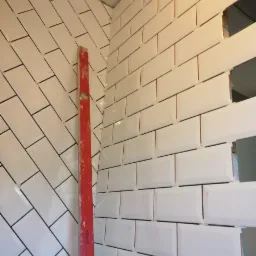 Układanie białych, błyszczących płytek w układzie cegiełki, widoczny poziomica i krzyżyki dystansowe. Częściowo wykończona ściana w rogu pomieszczenia.