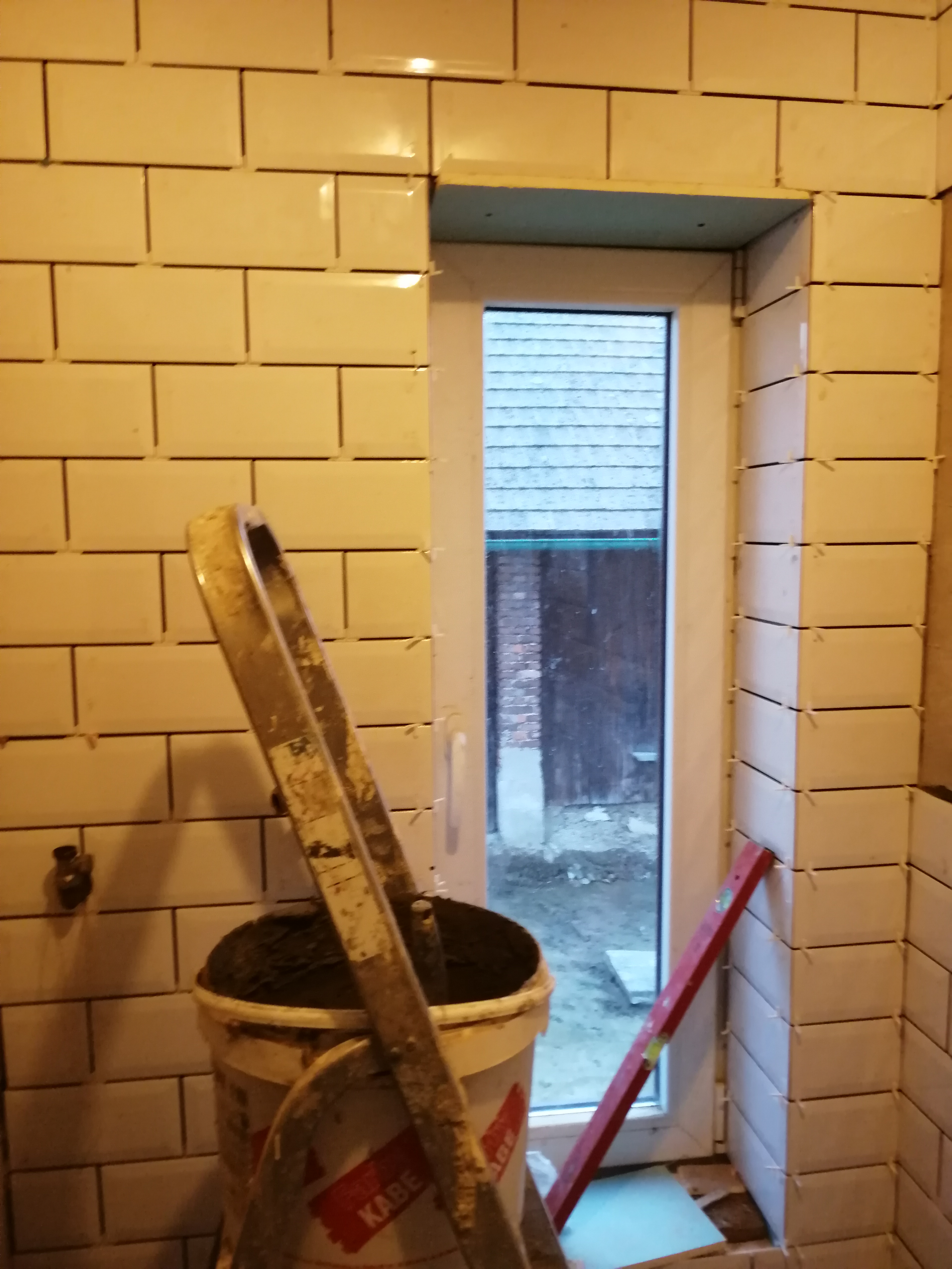 Ściana w trakcie układania białych, prostokątnych płytek, widoczna drabina oparta o ścianę i wiadro z zaprawą, poziomica przy oknie.