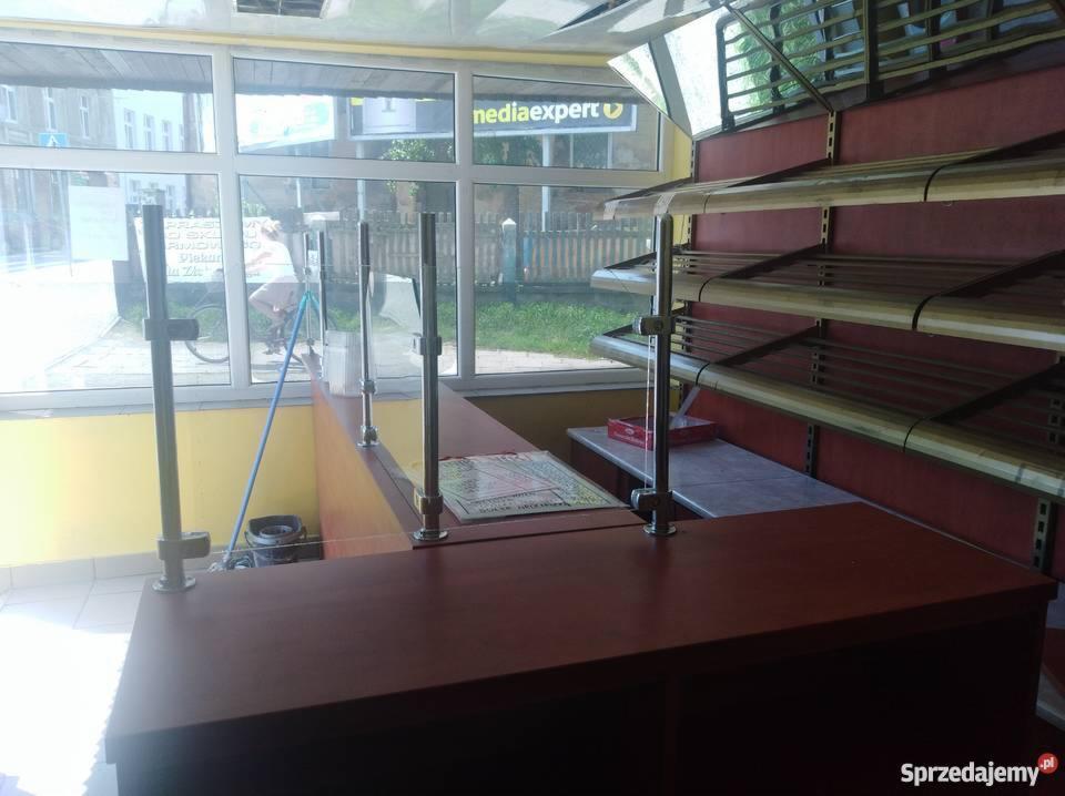 Pusta lada sklepowa z metalowymi przegrodami, regały bez towaru, widok na ulicę z rowerzystką za szybą.