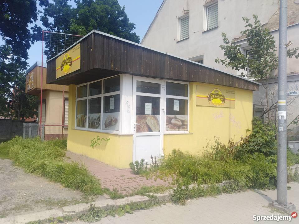 Narożny, parterowy budynek piekarni z żółtą elewacją, drewnianym daszkiem i reklamami pieczywa w oknach, otoczony zaniedbaną zielenią.