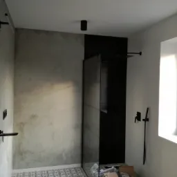 Nowoczesna łazienka w trakcie wykańczania: szara ściana z efektem betonu, kabina prysznicowa z czarnym panelem, wzorzysta posadzka, minimalistyczna armatura w czarnym macie, widoczne kartony...