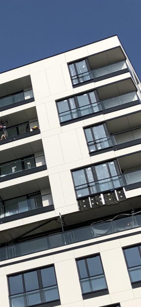Nowoczesny budynek w Warszawie z białą elewacją i czarnymi ramami okiennymi, częściowo w trakcie montażu izolacji na jednym z balkonów, widoczne rusztowania i pracownicy.