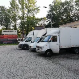 Trzy białe busy dostawcze zaparkowane na brukowanym placu przed budynkiem, z otwartymi drzwiami w pierwszym busie, widoczne drzewa w tle i szyld firmy CERAMTRANS II.