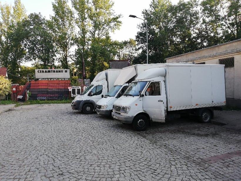 Trzy białe busy dostawcze zaparkowane na brukowanym placu przed budynkiem, z otwartymi drzwiami w pierwszym busie, widoczne drzewa w tle i szyld firmy CERAMTRANS II.