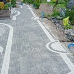 Ułożona szara kostka brukowa z białymi obrzeżami tworząca ścieżkę w ogrodzie, widoczne rośliny ozdobne i elementy architektury ogrodowej.