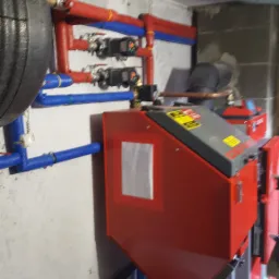 Czerwony piec z podajnikiem na ekogroszek, widoczne rury instalacji grzewczej w kolorze czerwonym i niebieskim, szara rura wentylacyjna pod sufitem, kotłownia.