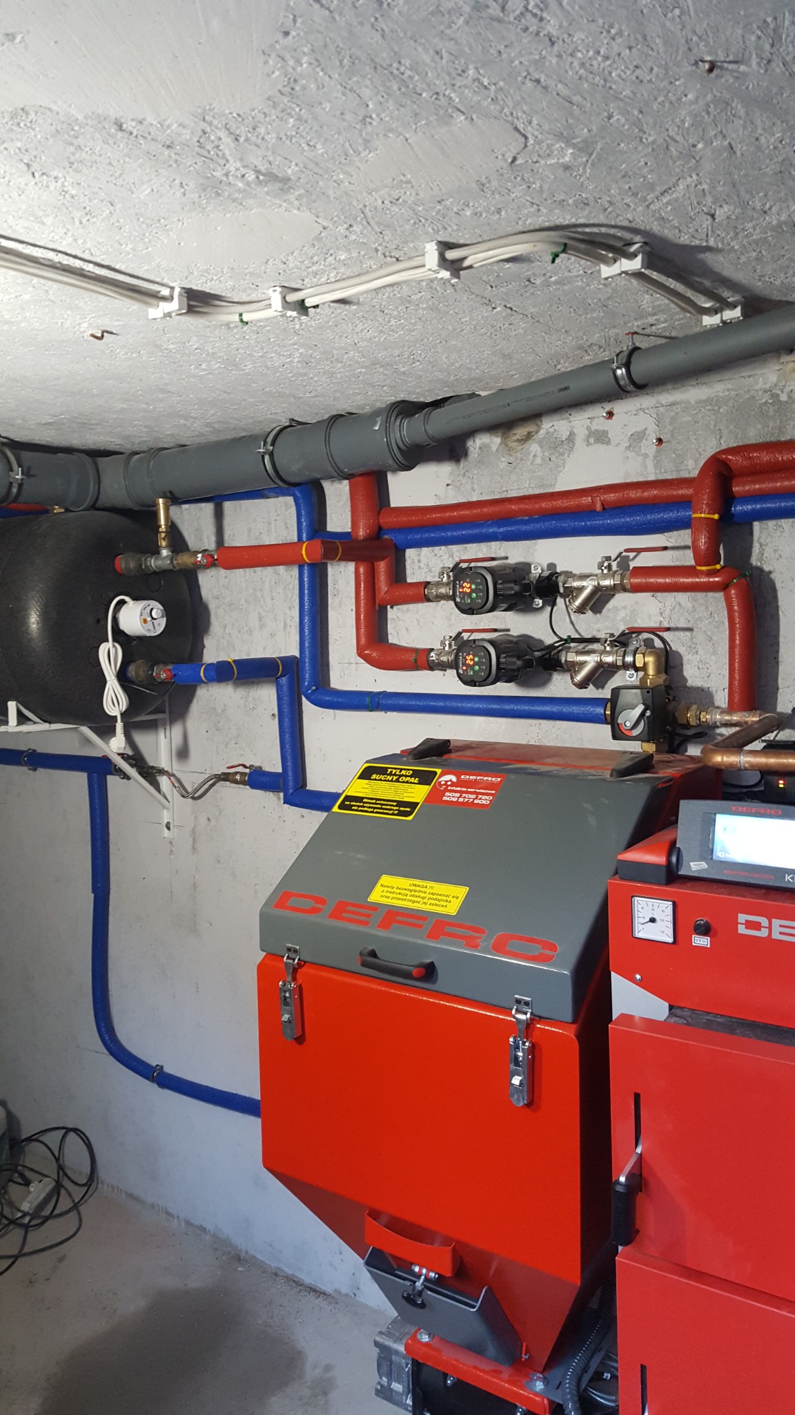 Kotłownia z piecem na paliwo stałe marki Defro, z instalacją grzewczą w rurach w kolorze czerwonym i niebieskim, zbiornikiem wyrównawczym i pompami obiegowymi, widoczne przewody elektryczne...
