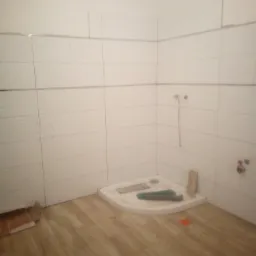 Remont łazienki w trakcie: białe płytki ścienne z poziomym wzorem, drewnopodobne panele podłogowe, brodzik w rogu pomieszczenia, widoczne przewody instalacji wodnej.